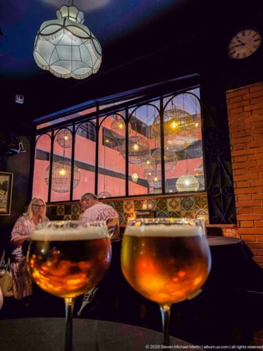 Two amber drinks in front of a decorated dining room with hanging lamps and seated patrons.