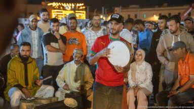 Man playing a large frame drum in a tight circle of seated musicians and standing spectators at night.