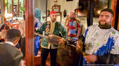 Three male musicians performing indoors, including a man in a red fez holding a large hand drum.
