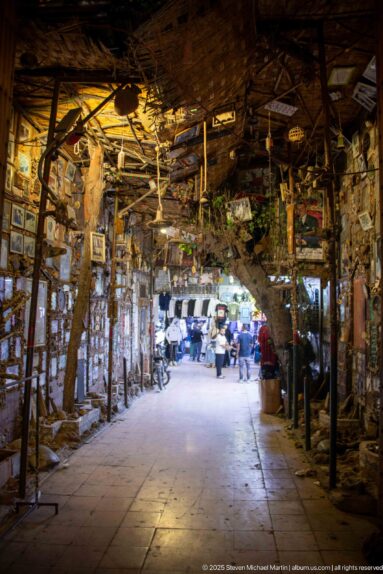 Rustic covered passage lined with framed pictures, hanging objects, and exposed overhead branches.