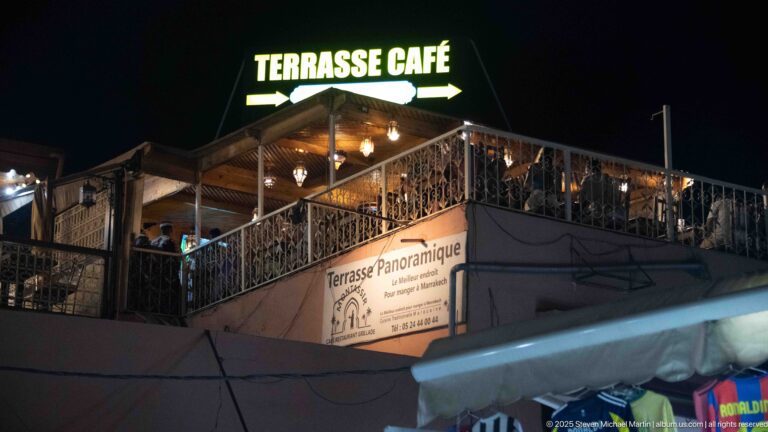 Rooftop café terrace at night beneath a bright TERRASSE CAFÉ sign and decorative railings.