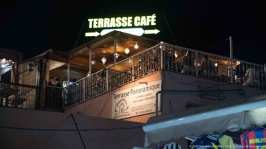 Rooftop café terrace at night beneath a bright TERRASSE CAFÉ sign and decorative railings.