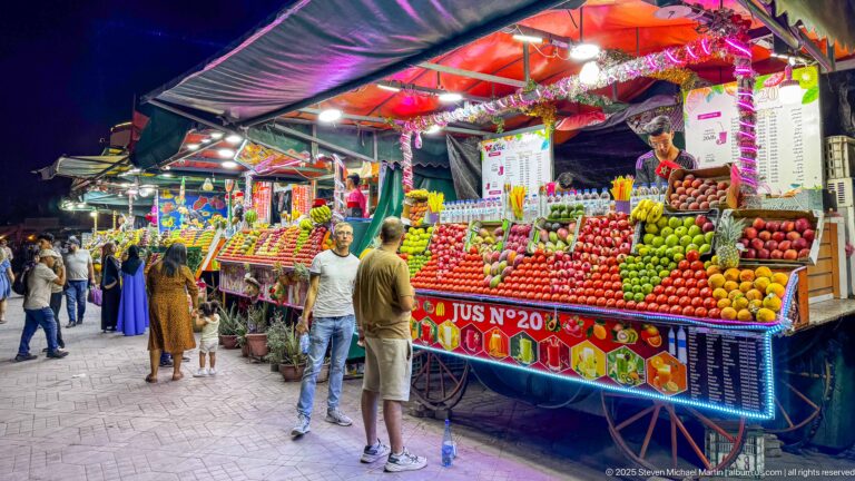 Bright juice stall No. 20 piled with colorful fruit as two customers stand at the counter in Jemaa el-Fna.