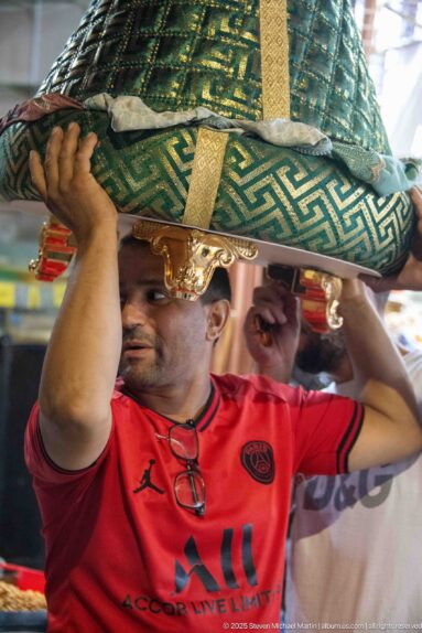 Man in a red shirt lifting a large green-and-gold patterned cushion or padded textile overhead.