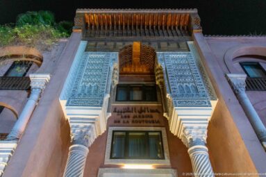 Ornate façade with carved plaster, twisted columns, wooden canopy, and bilingual sign above a doorway.