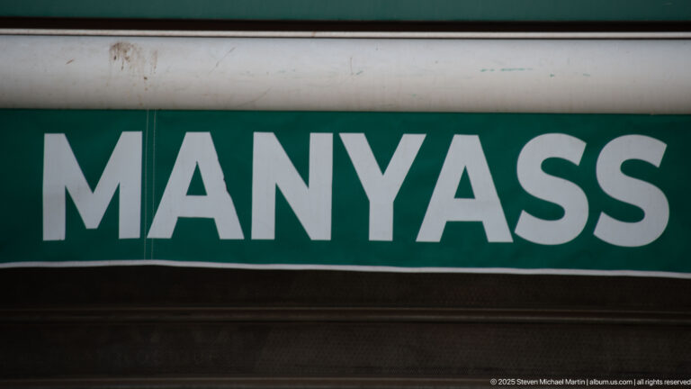 Close view of a green rectangular sign with white capital letters reading MANYASS.