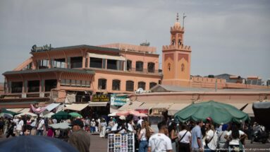 Busy market scene with crowds and umbrellas in front of low buildings and a square minaret.