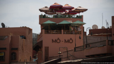 Rose-colored MÔ-MÔ building with rooftop terrace, patterned parasols, and green umbrellas.