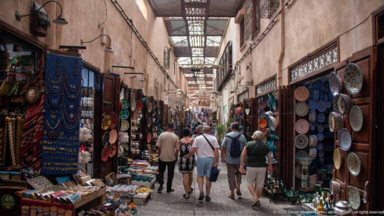 Covered souq lane with visitors walking between shops displaying plates, textiles, and crafts.