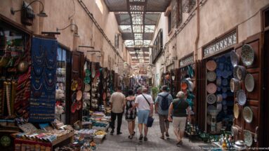 Covered souq lane with visitors walking between shops displaying plates, textiles, and crafts.
