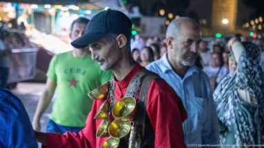 Street performer in a cap carrying several large brass-colored metal castanets across his chest in a nighttime crowd.