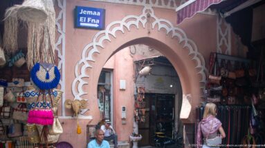 Pink scalloped arch with white trim opening into a narrow souq passage lined with hanging crafts.