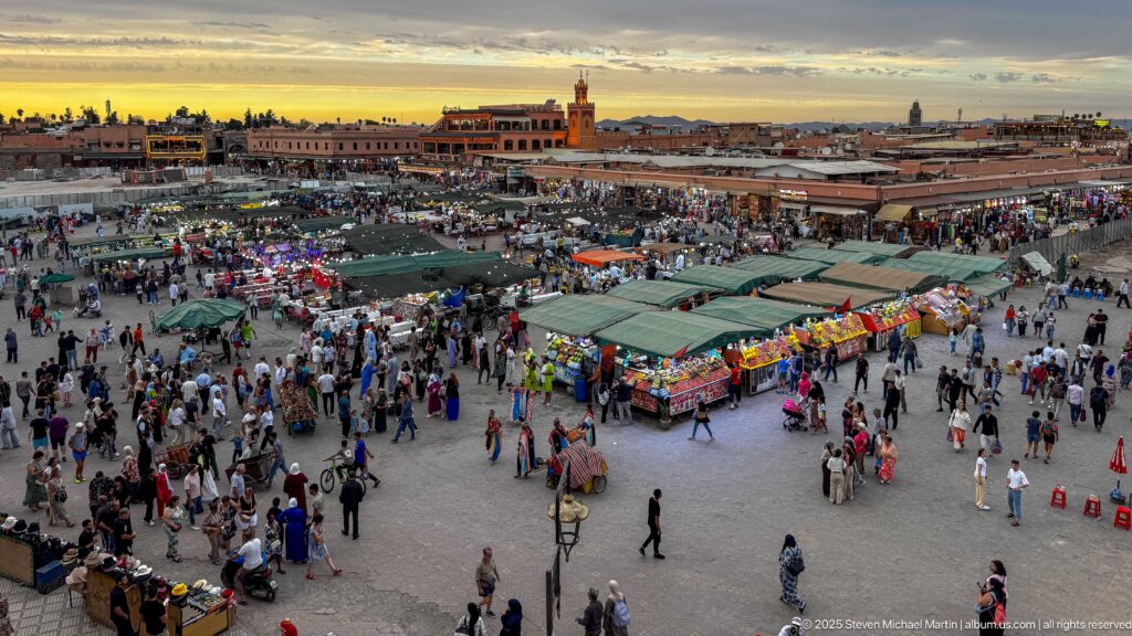 Elevated sunset panorama over Djemaa El Fna with food stalls, crowds, awnings, and low medina buildings.