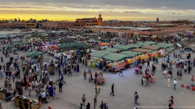Elevated sunset panorama over Djemaa El Fna with food stalls, crowds, awnings, and low medina buildings.
