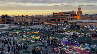 Evening view of crowded, brightly lit Djemaa El Fna food stalls with a lit tower beyond.