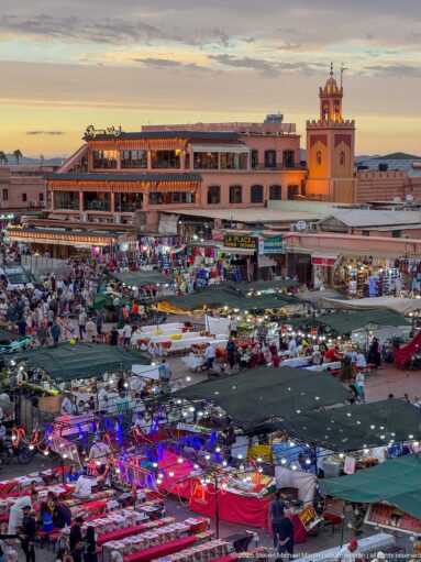 Market canopies and colorful goods across Djemaa El Fna with restaurants and an illuminated tower behind.