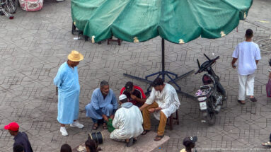 Men seated and standing beneath a large green umbrella on the paving of Djemaa El Fna.