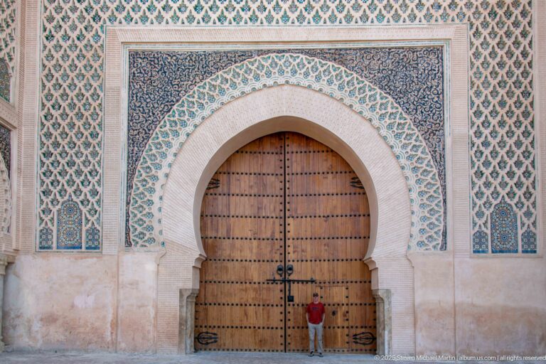 Large studded wooden double doors framed by green-and-blue geometric ornament, with a person standing below.