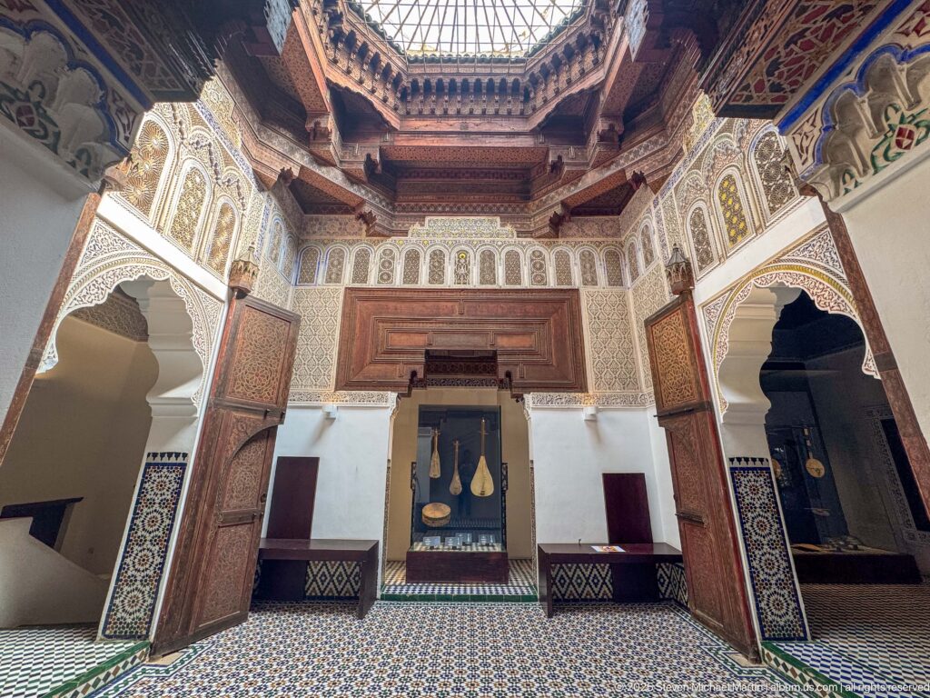 Wide ornate hall with open arches, zellij walls, carved woodwork, central display, and glazed ceiling lantern.