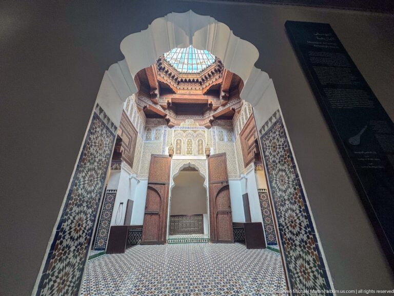 Low-angle view through a dark scalloped arch into a brightly decorated hall with tiled walls and ceiling lantern.