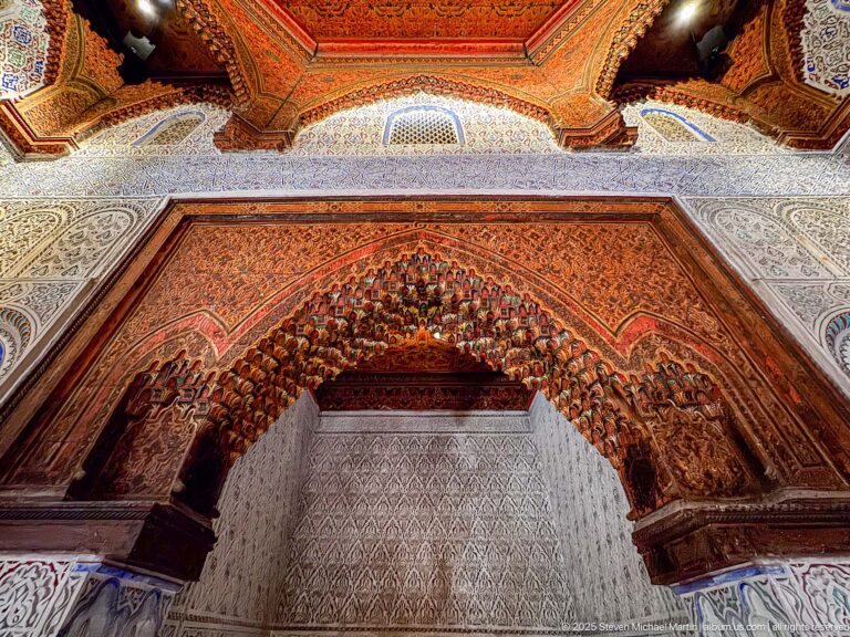 Close upward view of a richly painted muqarnas arch with layered carved niches in red, ochre, blue, and green.