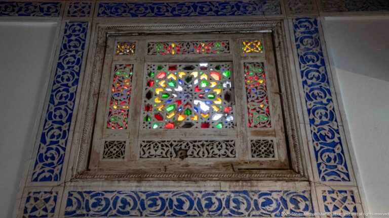 Small carved window with multicolored geometric stained glass, narrow side panels, and blue decorative borders.