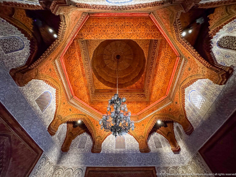 Upward view of an octagonal carved wooden ceiling with circular center, chandelier, arches, and small windows.