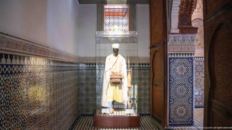 Mannequin in a long white robe with pale yellow layer and brown shoulder bag beside zellij walls.