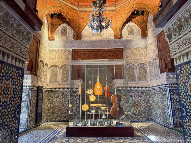 Glass case of traditional string instruments centered in a gallery with zellij, carved plaster, and wooden ceiling.