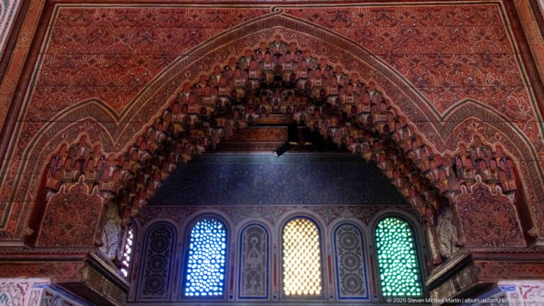 Carved red-brown muqarnas arch above narrow blue, amber, and green colored-glass windows.