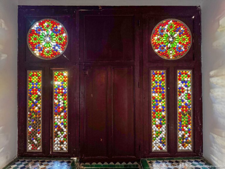 Dark wooden doors with two circular multicolored stained-glass rosettes above four narrow colored-glass panels.
