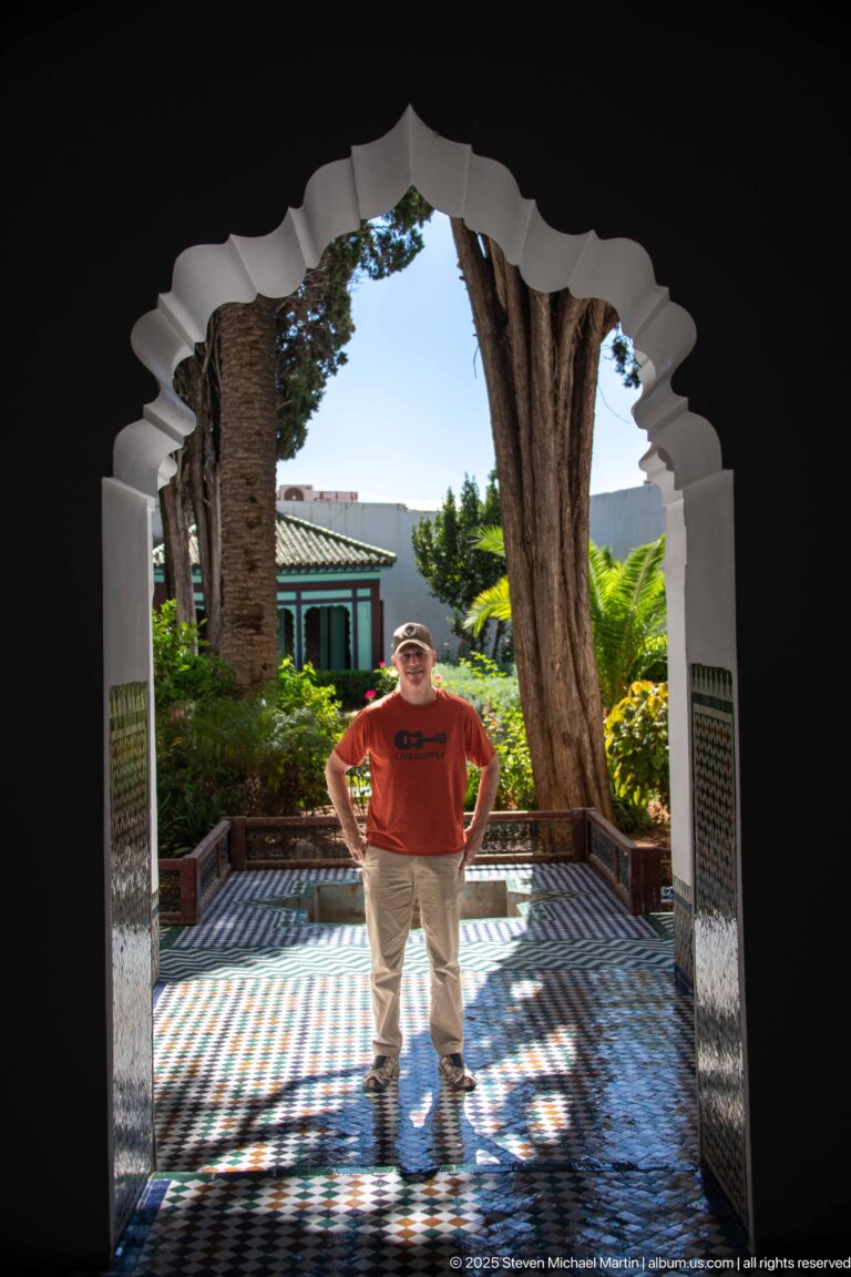 Visitor standing beneath a scalloped arch on patterned tile paving with trees and garden foliage behind.