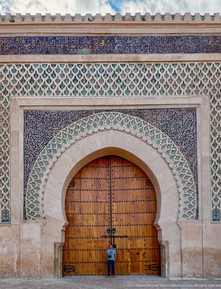 Frontal view of studded wooden doors beneath a pale arch and dense green geometric wall decoration.