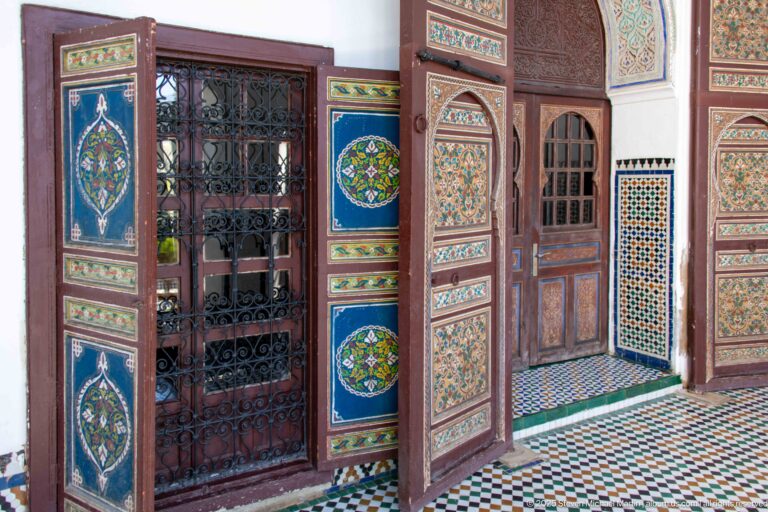 Open painted wooden doors with blue medallions framing a wrought-iron screen amid zellij and patterned floor tiles.