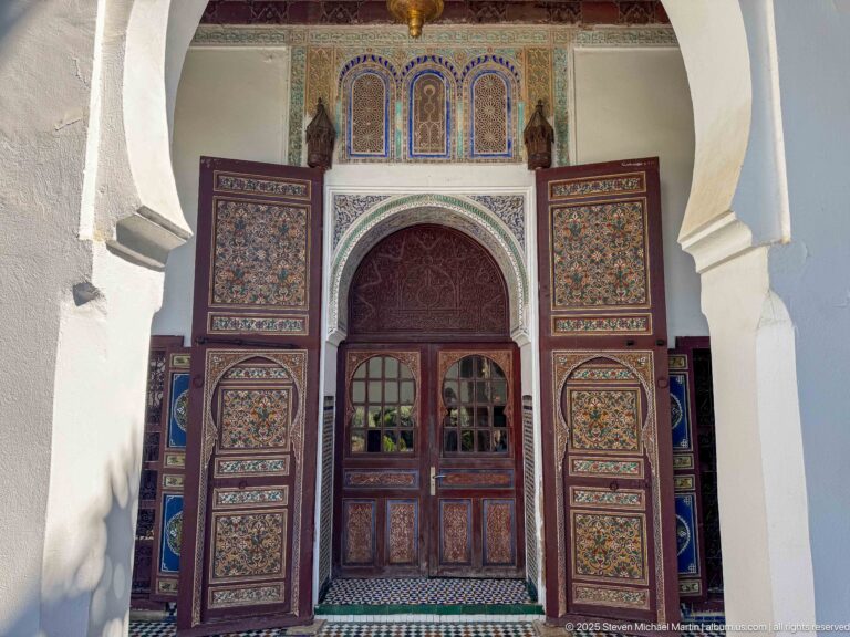 Painted wooden doorway beneath a decorated horseshoe arch, with open patterned shutters and three small panels above.