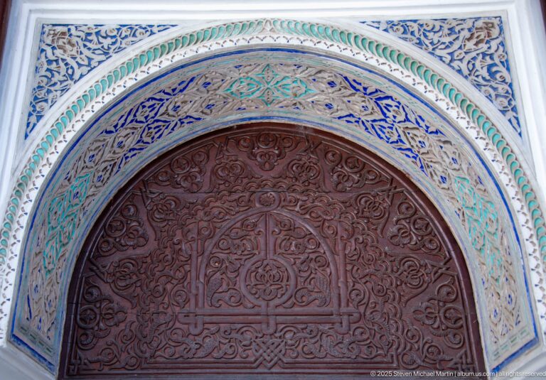Reddish carved wooden door beneath a horseshoe arch edged with blue, green, and white ornament.