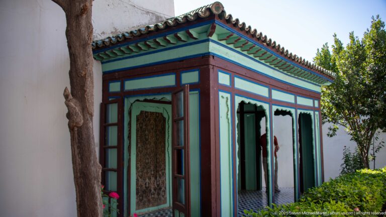 Small turquoise and burgundy painted garden pavilion with green-tiled roof beside trees and shrubs.