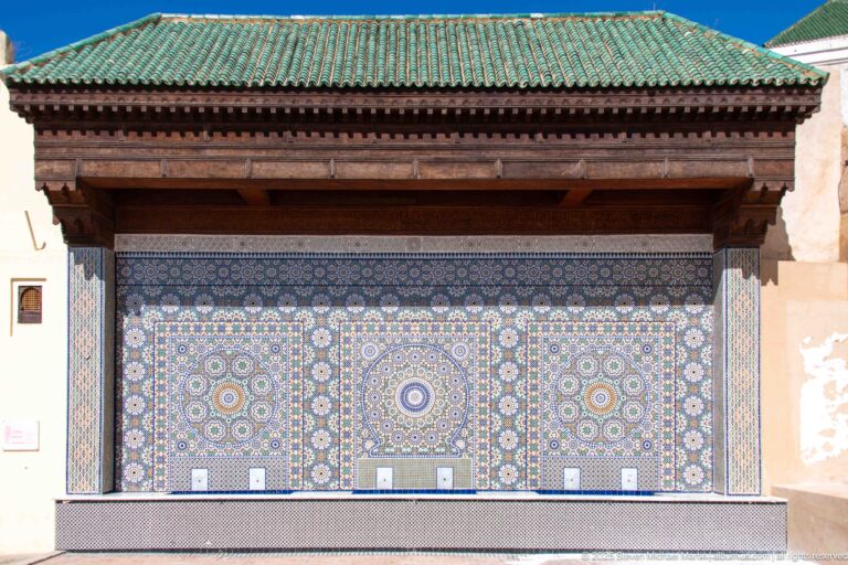 Large three-panel zellij wall fountain with radial rosettes beneath a carved wooden canopy and green-tiled roof.