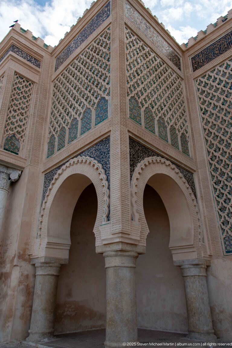 Oblique view of two horseshoe arches beneath dense green geometric panels and carved borders.