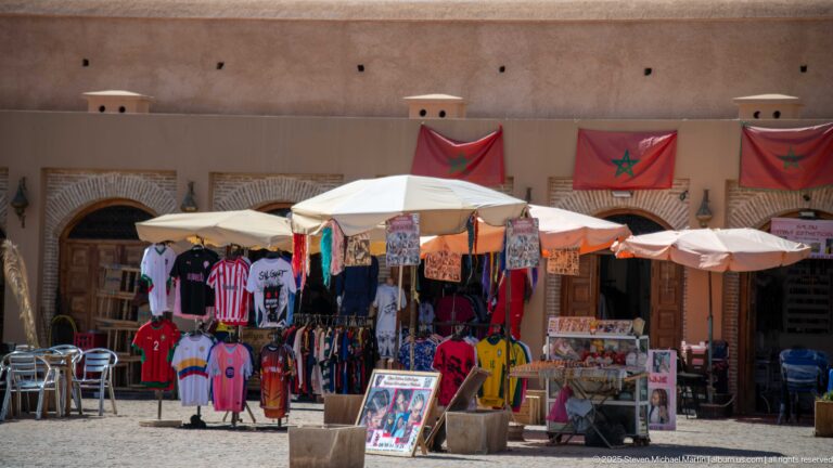 Market stall selling clothing and souvenirs beneath white umbrellas and Moroccan flags in front of arched wooden shop doors.