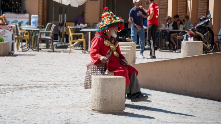 Seated Moroccan water seller in red clothing and colorful hat with metal cups and leather water bag.
