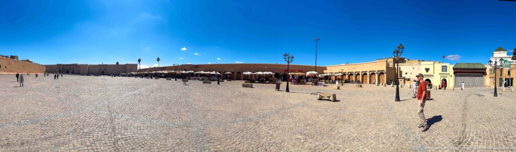 Wide paved square with scattered pedestrians, ochre walls, café umbrellas, and low buildings beneath a deep blue sky.