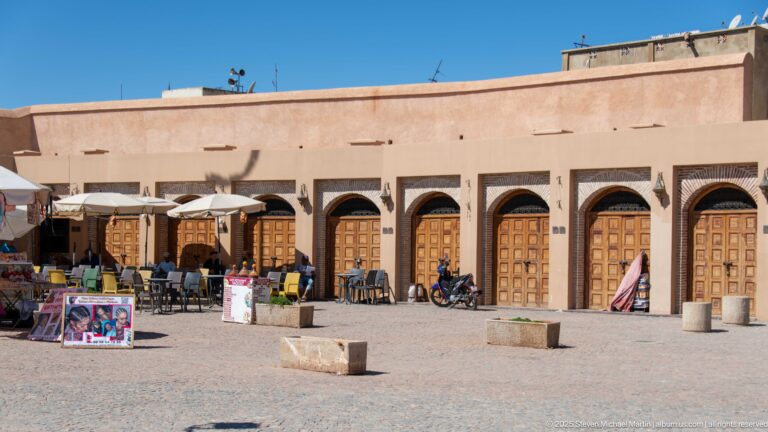 Row of paneled wooden shop doors beneath pointed arches, with café tables, umbrellas, signs, and merchandise outside.