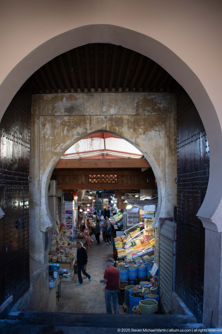Large arch opening into a covered produce market with vendors, shoppers, tubs, and crowded stalls.