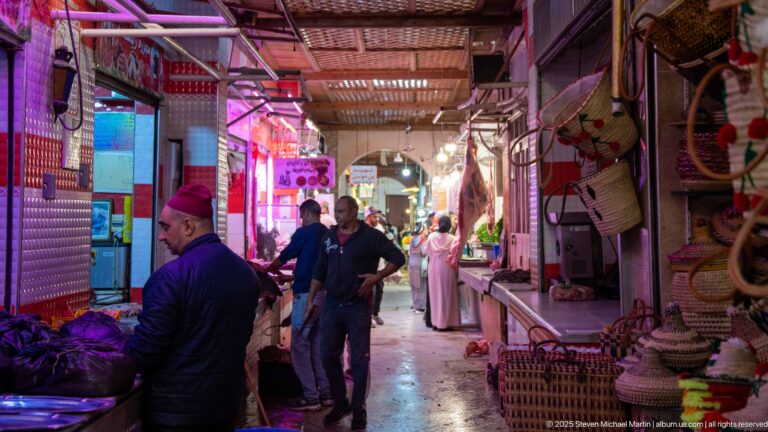 Narrow covered market aisle with vendors, shoppers, produce, hanging baskets, and woven household goods.