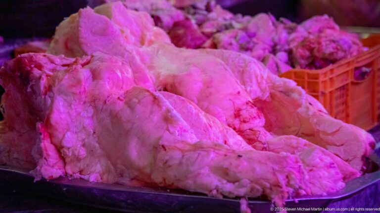 Close view of large pale raw meat cuts piled on a butcher counter under pink market lighting.