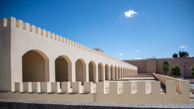 Long white arcade of rounded arches beneath crenellations, with repeating low barriers and a fortified wall beyond.