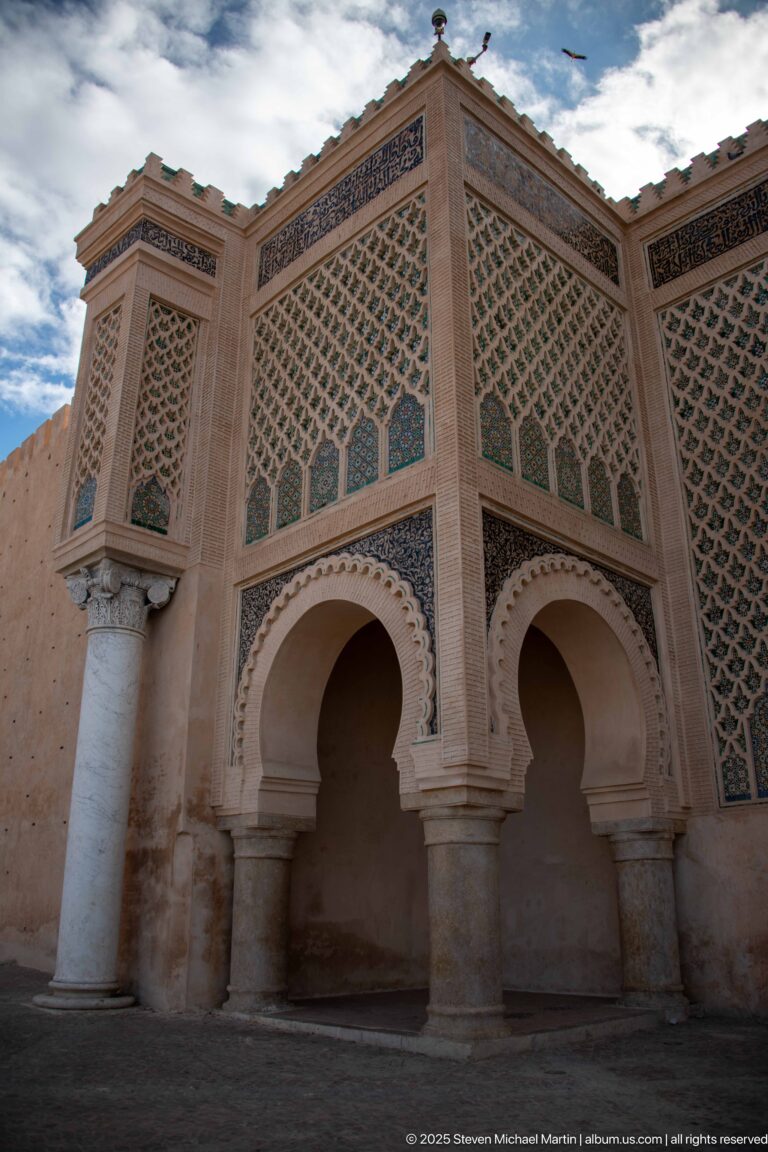 Low-angle corner view of two arches, columns, and tall patterned panels beneath a blue sky.