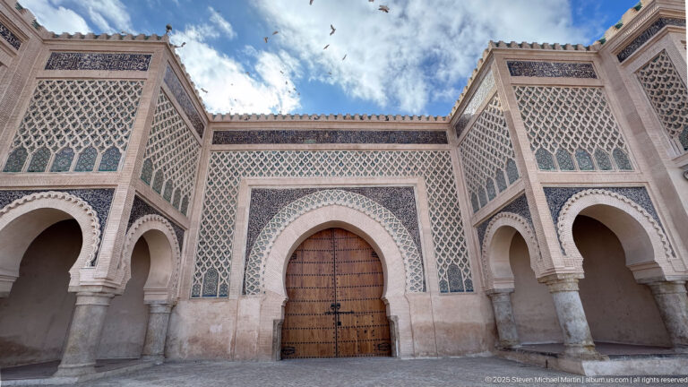 Wide frontal view of a decorated central gateway flanked by matching arches, with birds in the sky.