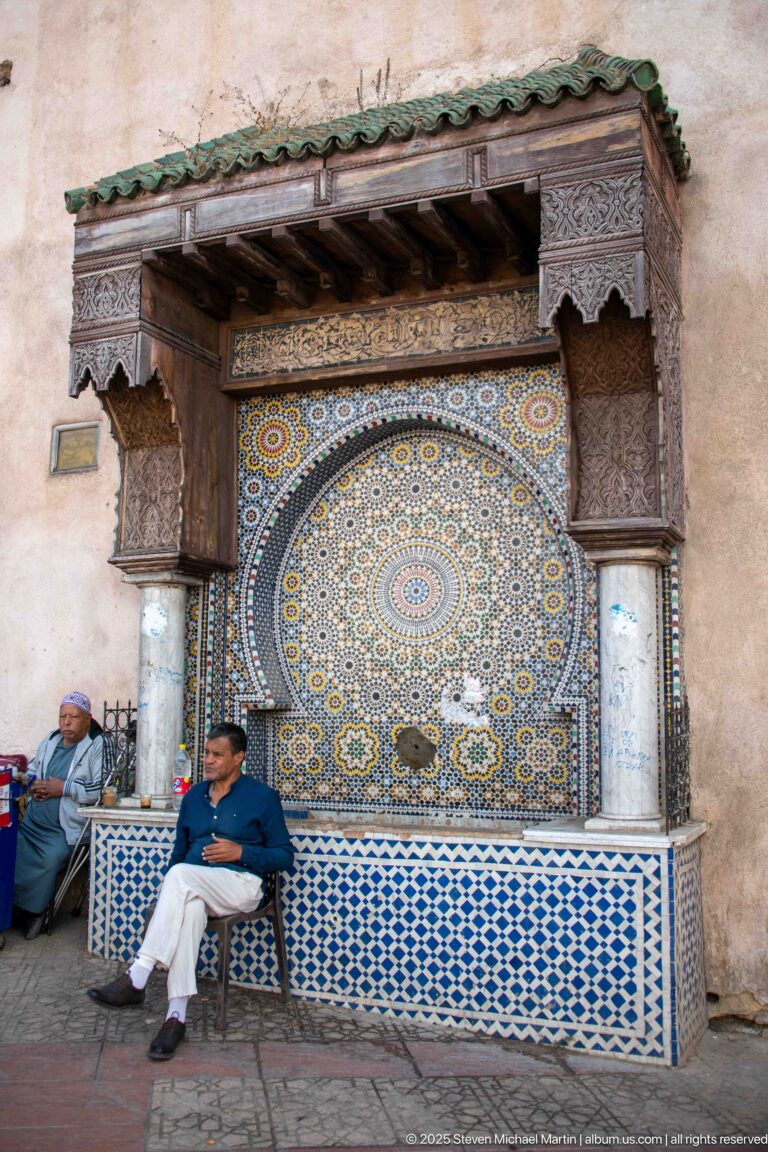 Decorative tiled wall fountain with a large circular mosaic rosette, flanked by columns and seated men.